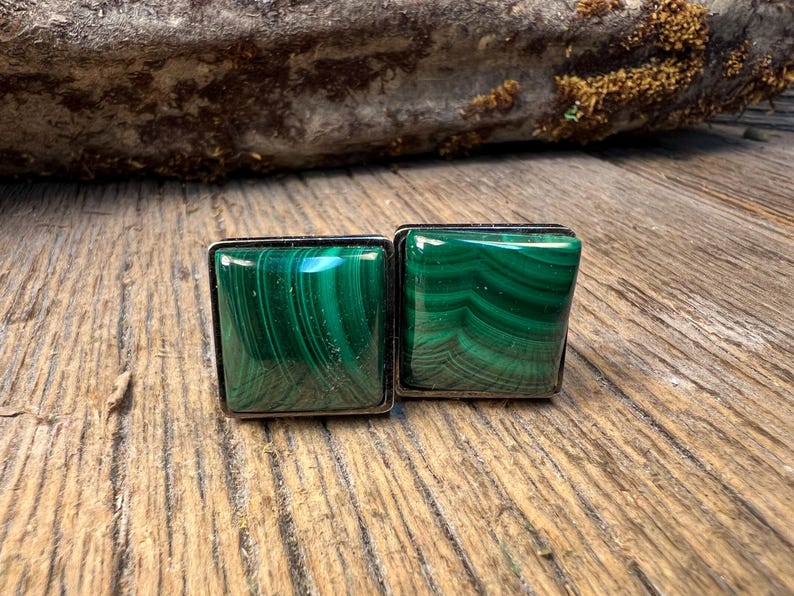 May include: Two square cufflinks with a silver border. The cufflinks are made of polished malachite, displaying a vibrant green color with swirling patterns. The cufflinks are displayed on a wooden surface.