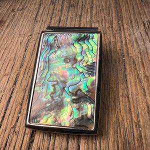 May include: A rectangular money clip featuring a polished metal frame and a central inlay of iridescent abalone shell. The shell displays a swirling, multi-colored pattern with vibrant shades of green, blue, pink, and purple that shift in the light.