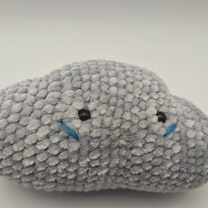 Peut inclure: Une peluche grise en forme de nuage avec une surface douce et texturée. Elle présente des yeux noirs en forme de bouton et des détails bleus cousus, créant un visage amical. Le jouet est conçu pour ressembler à un nuage duveteux.
