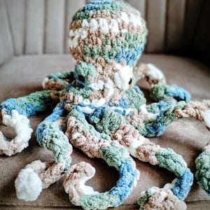 May include: A plush octopus toy crafted from variegated yarn in shades of blue, green, brown, and white. The stuffed animal features a rounded head and eight curled tentacles, with small black circular eyes positioned on the sides of the head.