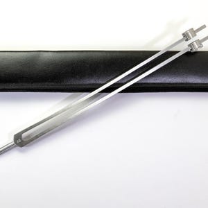 May include: A silver tuning fork with a long, slender handle and two prongs, resting on a black leather case. The tuning fork is designed for producing a specific musical pitch. The case is rectangular and has a zipper closure.