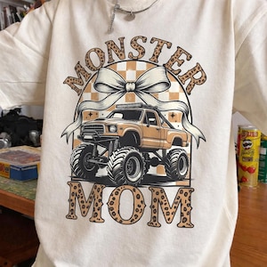 Op de afbeelding: Crèmekleurige sweatshirt met een afbeelding van een monstertruck met grote banden, een strik en de woorden "MONSTER MOM" in een luipaardprint lettertype. De achtergrond bevat een dambordpatroon.