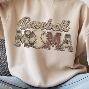 Puede incluir: Una sudadera beige con las palabras "Baseball MOMA" en un diseño cosido. La palabra "Baseball" está en una fuente marrón texturizada, y la pelota de béisbol está en el centro. La palabra "MOMA" está en un patrón de camuflaje.