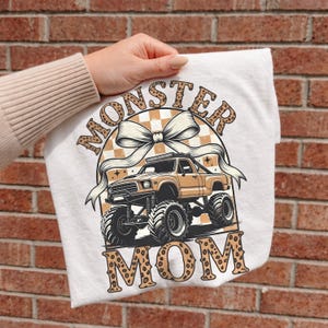 Op de afbeelding: Wit T-shirt met een bruin monster truck-ontwerp en de woorden "MONSTER MOM". Het ontwerp bevat een strik en een geruite achtergrond. Het shirt wordt door een persoon vastgehouden.