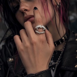 May include: A silver-colored ring shaped like a set of fangs. The ring has sharp, pointed teeth and is worn on a finger. The ring is a unique piece of jewelry.