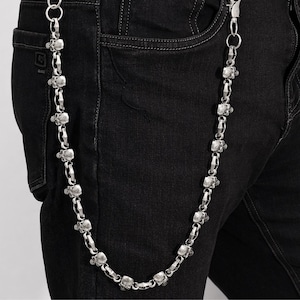 Puede incluir: Una cadena de billetera plateada con eslabones en forma de calavera. La cadena está sujeta a unos vaqueros negros. La cadena es un accesorio decorativo.