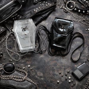 Puede incluir: Dos bolsos cruzados con forma de ataúd, uno transparente y otro negro, con adornos de arañas. Los bolsos se exhiben con cadenas, una chaqueta de cuero con tachuelas y otros accesorios góticos sobre una superficie oscura y texturizada.