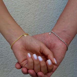 May include: Two wrists are shown, each adorned with a delicate bracelet. One is gold with a 'B' charm, the other silver with an 'E' charm. The bracelets have a rope chain design, adding a touch of elegance.
