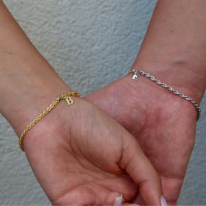 May include: Two rope chain bracelets, one gold and one silver, each with a single initial charm. The gold bracelet has a 'B' charm, and the silver bracelet has an 'E' charm. The bracelets are worn on wrists.