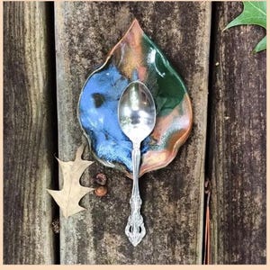 May include: A silver spoon rests on a leaf-shaped ceramic dish with blue, green, and gold glaze. The dish sits on a weathered wooden surface, with a dried leaf and small acorns nearby. The spoon has an ornate handle.