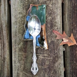 May include: A silver-toned metal spoon with an ornate handle resting on a ceramic spoon rest shaped like the state of Mississippi. The ceramic piece features a glossy glaze with blended shades of deep green, brown, and blue along the edges.