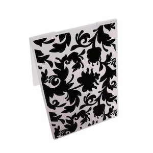 May include: A white compact mirror with a black floral design. The pattern features stylized flowers and leaves. The mirror is rectangular and has a hinged design.