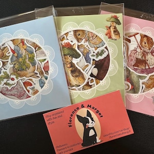 Puede incluir: Tres paquetes de pegatinas en azul, verde y rosa, cada uno con un diseño circular de conejos ilustrados y otros personajes. Una tarjeta de visita con un logotipo de perro en blanco y negro está al frente. La tarjeta dice "Florence & Margot".