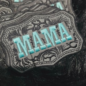 May include: A stack of rectangular embroidered patches featuring a gray background with black decorative scrollwork borders. The word MAMA is stitched in the center using bright turquoise thread with a bold, block-style font and black outlines.