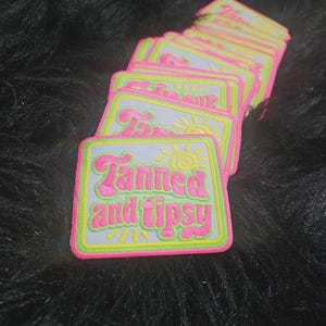 Könnte beinhalten: Ein Stapel rechteckiger bestickter Aufnäher mit weißem Hintergrund und neonpinkem Rand. Der Text Tanned and tipsy ist in Neonpink gestickt, mit einem neongelben Sonnensymbol über dem Text und gelben Strahlen unter dem Text.