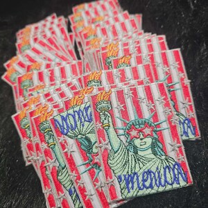 May include: A stack of rectangular embroidered patches featuring the Statue of Liberty wearing star-shaped red glasses. The design includes a background of red and white stripes with silver stars and the word 'merica' in blue script embroidery.