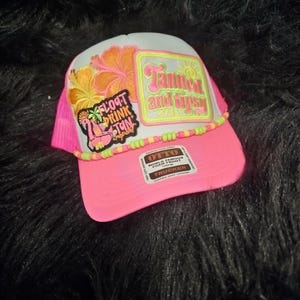 May include: A bright pink trucker hat featuring a white front panel with a patch that reads Tanned and Tipsy and a secondary patch with a flamingo and the text Float Drink Tan Repeat. The hat is accented with a string of yellow and pink beads.