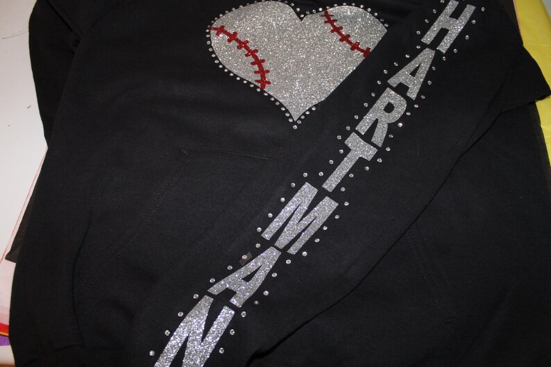 May include: Black hoodie with a silver glitter heart-shaped baseball design and the word "HEART" spelled out in silver glitter on the sleeve.