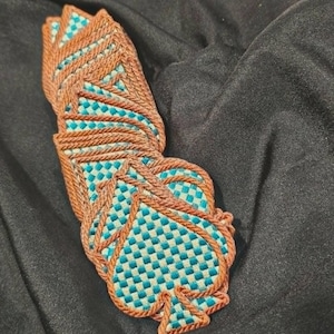 May include: A stack of spade-shaped coasters featuring a woven checkerboard pattern in teal and light blue, bordered by a thick, raised brown corded edge. The coasters are arranged in a slightly fanned, overlapping vertical column.