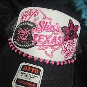 May include: A black and white trucker hat featuring a white foam front panel with pink and black embroidery. The design includes playing cards, a Texas state outline, and the text "She's from TEXAS I CAN TELL", "Raisin' Hell", and "Yee Haw".