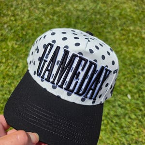 May include: A baseball cap featuring a white crown with a black polka dot pattern and a solid black brim. The front of the crown displays the word GAMEDAY in bold, black, embroidered, block-style lettering with a white outline.
