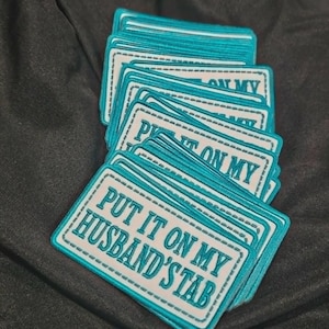 May include: A stack of rectangular embroidered patches with a white background and teal borders. The text "PUT IT ON MY HUSBAND'S TAB" is stitched in teal capital letters across the center of each patch, surrounded by a dashed teal rectangular border.