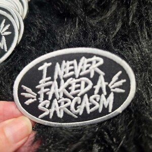 May include: An oval-shaped embroidered patch with a black background and a white border. The patch features the white embroidered text "I NEVER FAKED A SARCASM" in a stylized font, with small white star-like accents on the left and right sides.