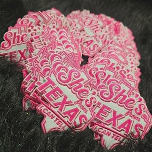May include: A collection of embroidered patches featuring a white background with bright pink stitched lettering that reads She's from TEXAS I CAN TELL. The patches are shaped like the state of Texas and are stacked together in a pile.