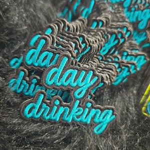 May include: A stack of embroidered patches featuring the text day drinking in bright turquoise thread with a dark brown border. The patches are layered on top of each other, showcasing the cursive script and the raised texture of the embroidery.