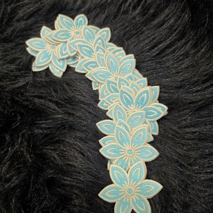 May include: A collection of multiple embroidered flower patches arranged in a curved line. Each patch features a six-petaled blossom design with a bright light blue center and a contrasting cream-colored stitched border outline.