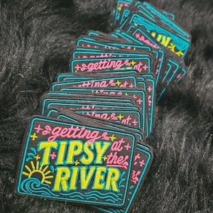 May include: A stack of rectangular embroidered patches with a black background and teal borders. The text reads "getting at the TIPSY RIVER" in pink and yellow thread. A yellow sun icon and teal wave graphics are visible on the left side of the patch.