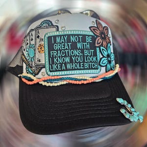 May include: Black trucker hat with a mesh back and a curved brim. The front panel features a patch with the text "I MAY NOT BE GREAT WITH FRACTIONS, BUT I KNOW YOU LOOK LIKE A WHOLE BITCH." Embellished with turquoise accents and floral designs.