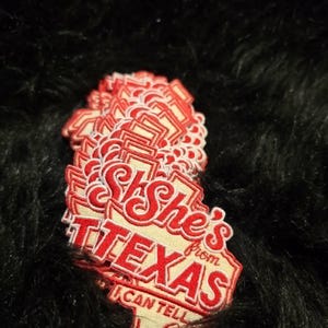 May include: A stack of embroidered patches shaped like the state of Texas. The patches feature a cream-colored background with red stitched borders and red text that reads: She's from TEXAS I CAN TELL.