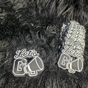 May include: Two black and white embroidered patches featuring the text Let's GO in a stylized script and block font, accompanied by a megaphone graphic. The patches have a thick white border and are designed for application to fabric surfaces.