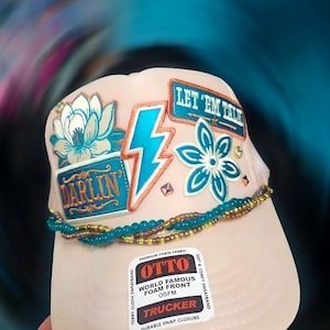 May include: A light beige trucker hat featuring a foam front adorned with various patches including a blue lightning bolt, a flower, and text reading "DARLIN'" and "LET 'EM TALK". The hat is decorated with strands of teal and gold beads.