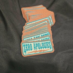May include: A stack of rectangular embroidered patches featuring a light-colored background with a teal border and the text ZERO APOLOGIES in teal lettering. Below the text is a small teal lightning bolt graphic, all framed by an orange stitched edge.
