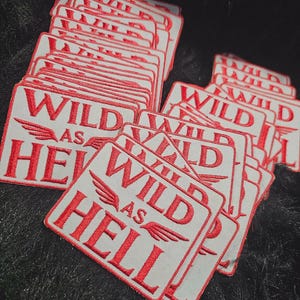 May include: A stack of rectangular embroidered patches featuring a white background with red borders and red text that reads WILD AS HELL, accompanied by a small winged graphic element positioned between the words WILD and AS.