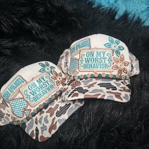 May include: Two trucker hats featuring a white front panel with a patch that reads "ZERO APOLOGIES" and "ON MY WORST BEHAVIOR" in teal lettering. The hats have a brown, white, and gray camouflage brim, floral accents, and a beaded trim across the base.