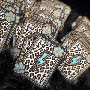 May include: A collection of embroidered patches shaped like playing cards, each featuring an ace of spades symbol, a leopard print pattern, a central light blue lightning bolt, and a light blue flower accent with black stitched borders.
