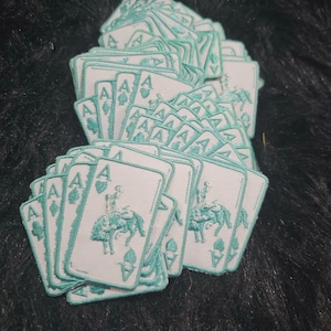 Könnte beinhalten: Ein Stapel rechteckiger Stickaufnäher, jeweils mit weißem Hintergrund, türkisfarbenem Rand und einer türkisfarbenen Grafik eines Cowboys auf einem bockenden Pferd. Jeder Aufnäher zeigt den Buchstaben A in den gegenüberliegenden Ecken.
