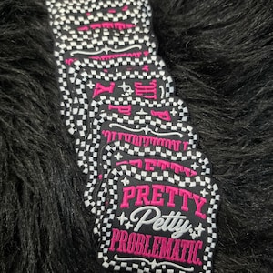 May include: A stack of square embroidered patches with scalloped edges featuring a black and white checkered border. The center is black with the pink embroidered text PRETTY, the white script text Petty, and the pink embroidered text PROBLEMATIC.