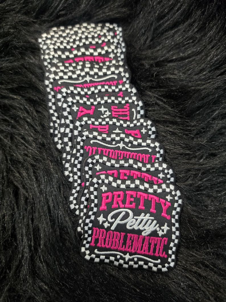 May include: A stack of square embroidered patches with scalloped edges featuring a black and white checkered border. The center is black with the pink embroidered text PRETTY, the white script text Petty, and the pink embroidered text PROBLEMATIC.