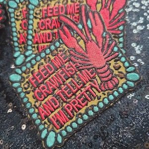 May include: A rectangular embroidered patch featuring a red crawfish illustration and the text FEED ME CRAWFISH AND TELL ME I'M PRETTY in red thread. The patch has a gold-colored background with a border of teal-colored circular shapes.