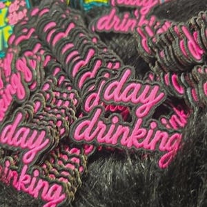 May include: A collection of embroidered patches featuring the text day drinking in bright pink cursive script with a black border. The patches are stacked together, showing the repeating design and the vibrant pink thread against the black fabric.