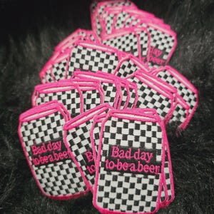 May include: A collection of embroidered patches shaped like beverage cans, featuring a black and white checkered pattern with a bright pink border. Each patch displays the text "Bad day to be a beer" in pink embroidery on a black rectangular background.