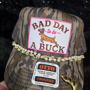 May include: A camouflage trucker hat featuring a rectangular patch with the text BAD DAY to be A BUCK and an embroidered deer. The hat is adorned with a gold-tone chain and pearl trim, a gold antler charm, and a sticker label reading OTTO TRUCKER.