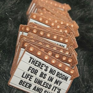 May include: A stack of embroidered patches shaped like a marquee sign with a brown border and light-colored center. The text reads: THERE'S NO ROOM FOR BS IN MY LIFE UNLESS IT'S BEER AND SHOTS. The design includes small circular accents along the top edge.