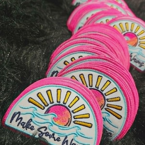 May include: A stack of semi-circular embroidered patches featuring a pink and orange gradient sun, yellow rays, and light blue waves. The text "Make Some Waves" is embroidered in dark blue script across the bottom of each white patch.