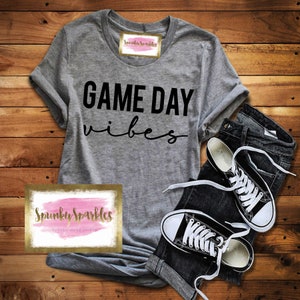 May include: A gray t-shirt with the words "GAME DAY vibes" in black script, paired with dark denim jeans and black and white sneakers. The shirt has a logo that says "Spunky Sparkles" in gold lettering.