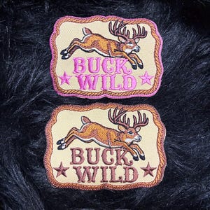 May include: Two rectangular embroidered patches with scalloped edges, each featuring a leaping deer and the text BUCK WILD flanked by two stars. The top patch has pink text and border, while the bottom patch has brown text and border on a tan background.